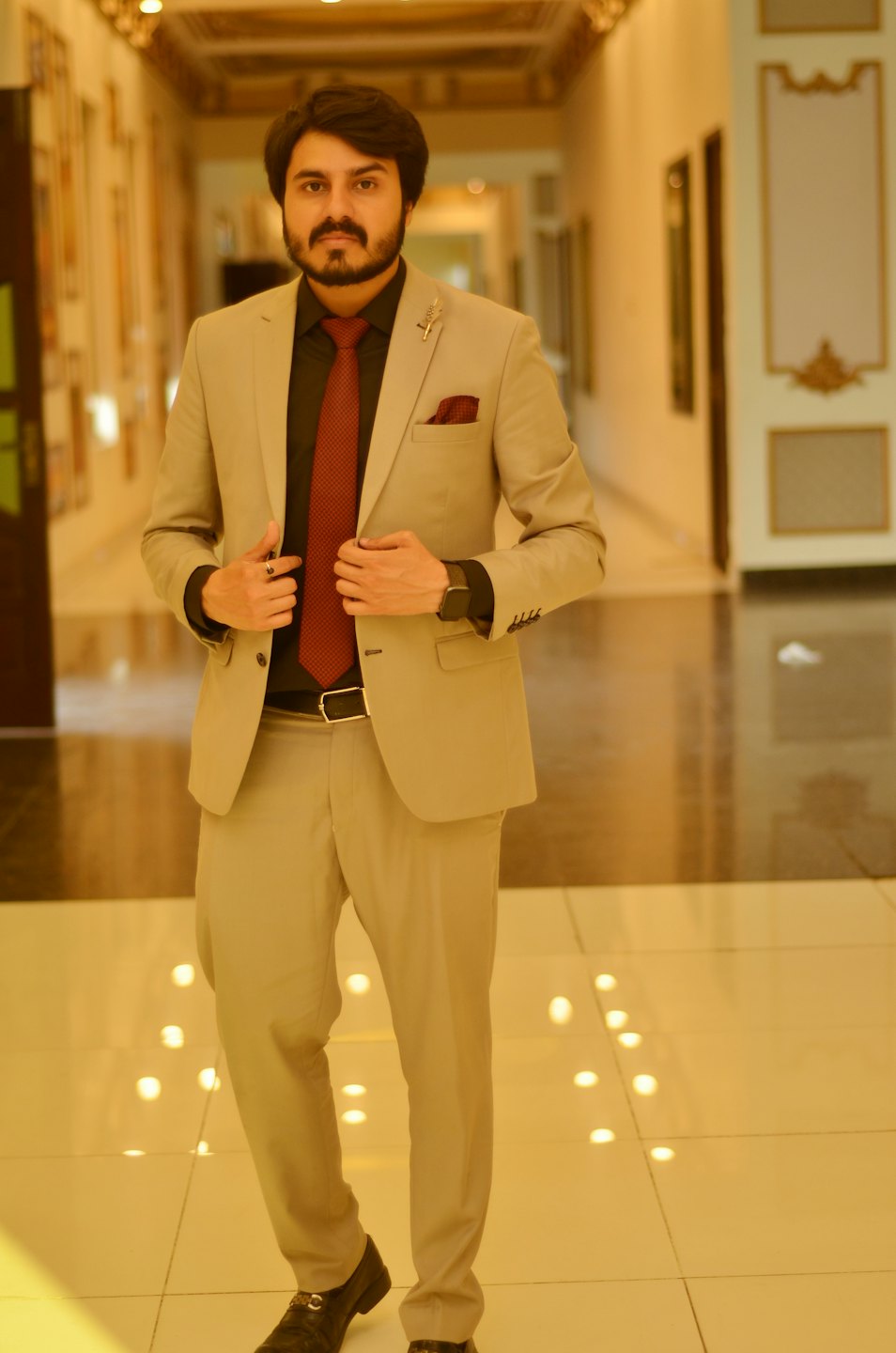 Man in a beige suit adjusts his lapels in a hallway.