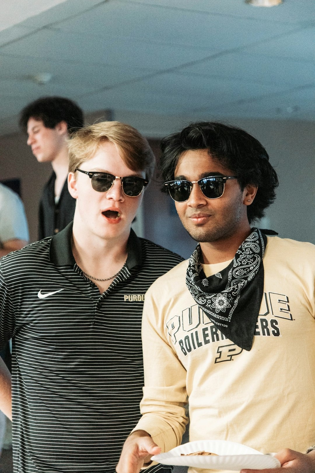 Two men wearing Purdue apparel hold food while standing indoors.
