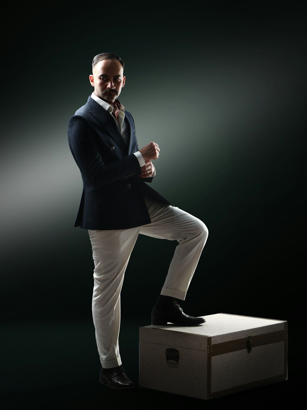 Man in a suit adjusts his cuff while resting his foot on a trunk.
